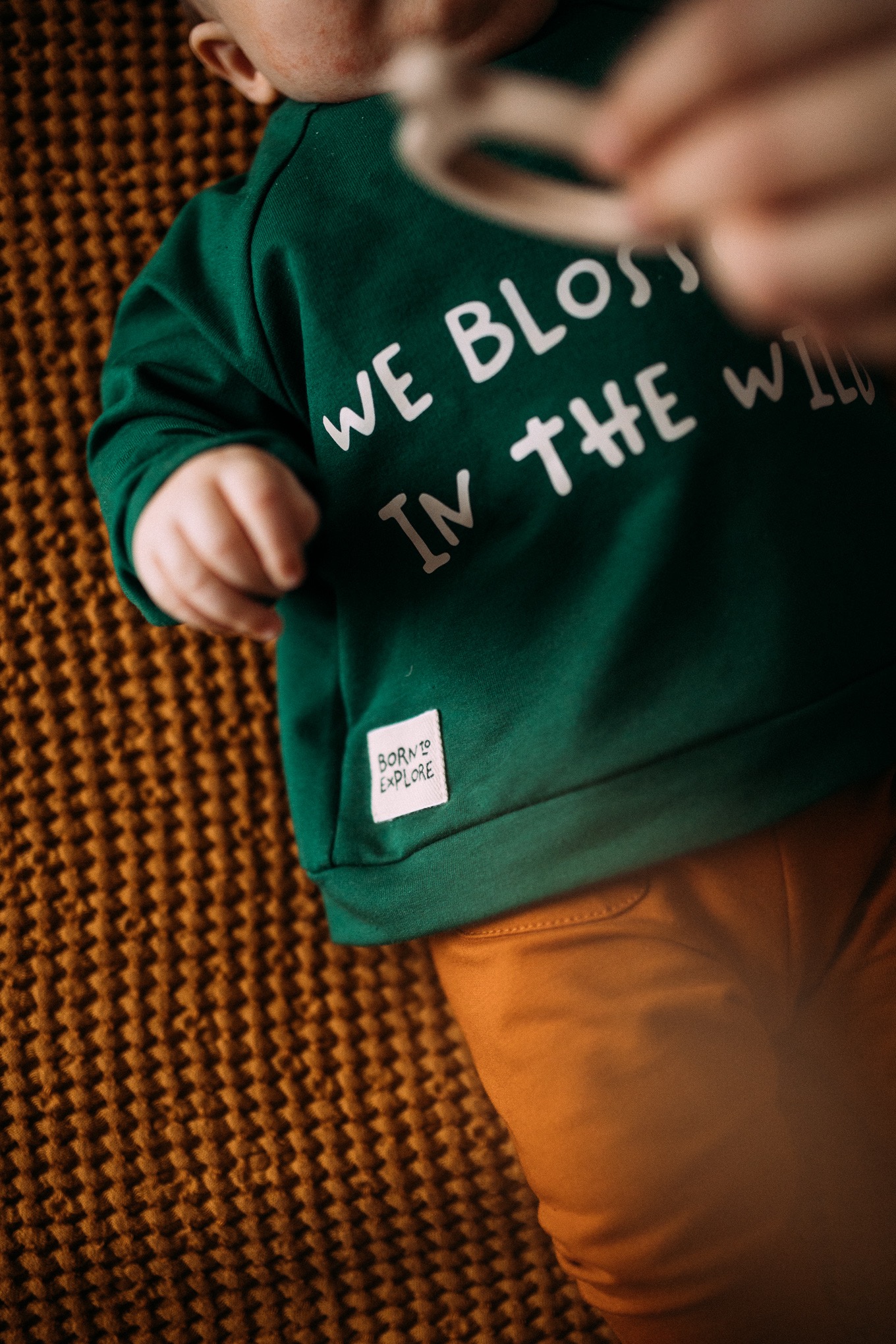 Niemowlę leżące na brązowym kocu w zielonej bluzie z napisem 'We blossom in the wild' i metką 'Born to explore', ubrane w pomarańczowe spodnie.