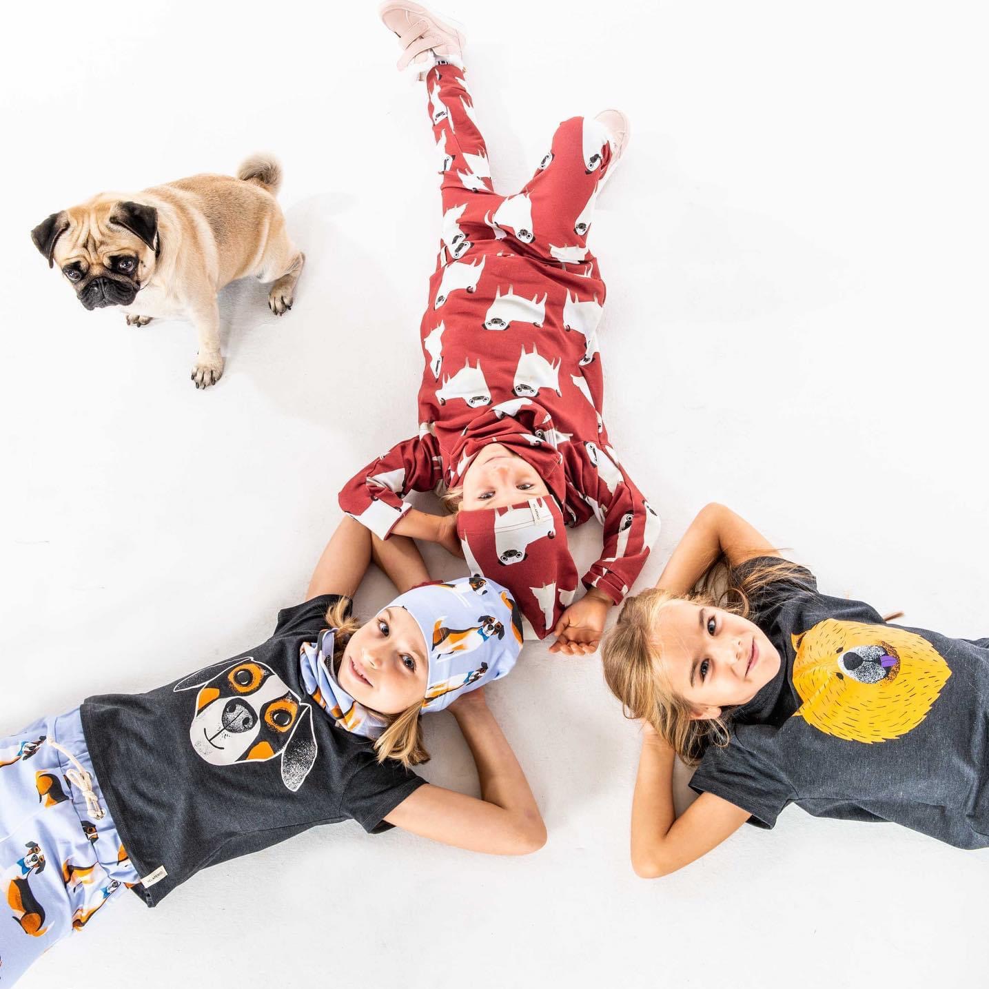 Trzy dziewczynki leżące na białym tle w ubraniach z nadrukami zwierząt, obok stoi mops patrzący w górę.