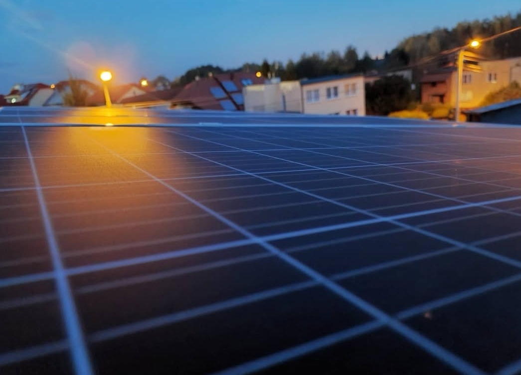 Instalacja paneli słonecznych na dachu budynku, widoczna wieczorową porą pod światłem latarni, zarys budynków w tle, równomierne rozłożenie modułów.