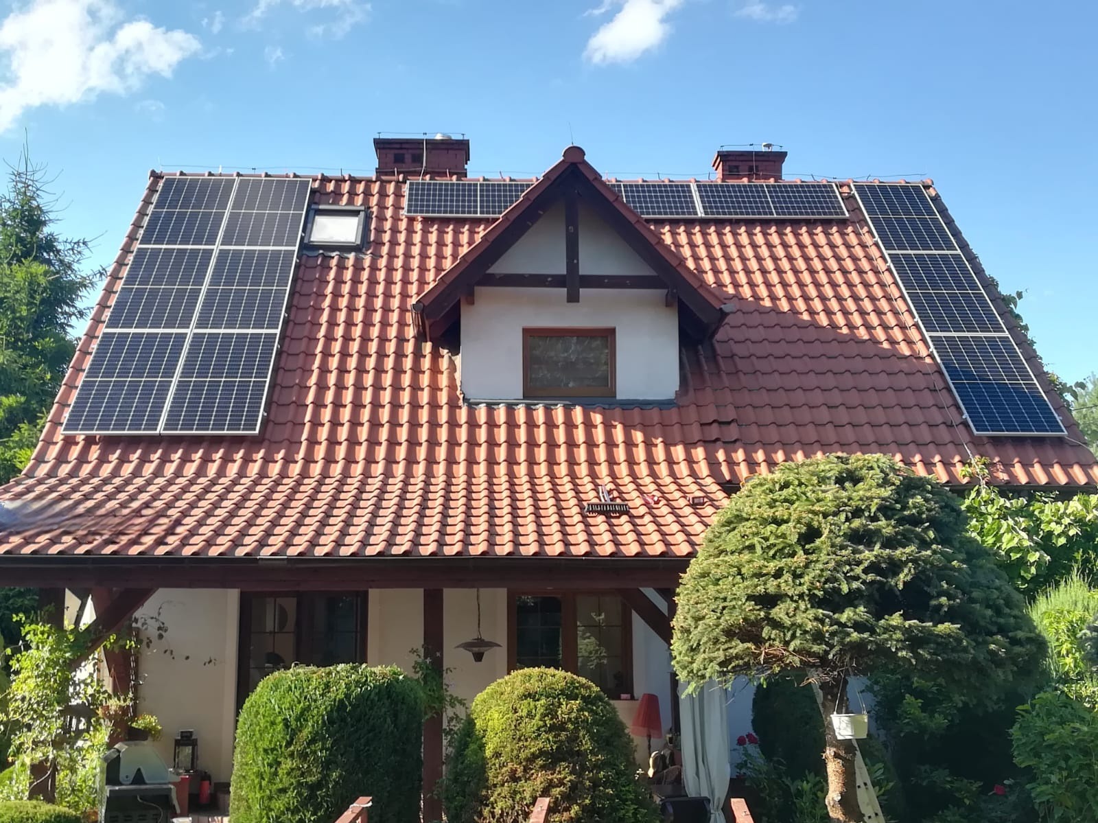 Dom jednorodzinny z panelami słonecznymi na dachu z czerwonej dachówki. Widok z poziomu ogrodu, błękitne niebo.