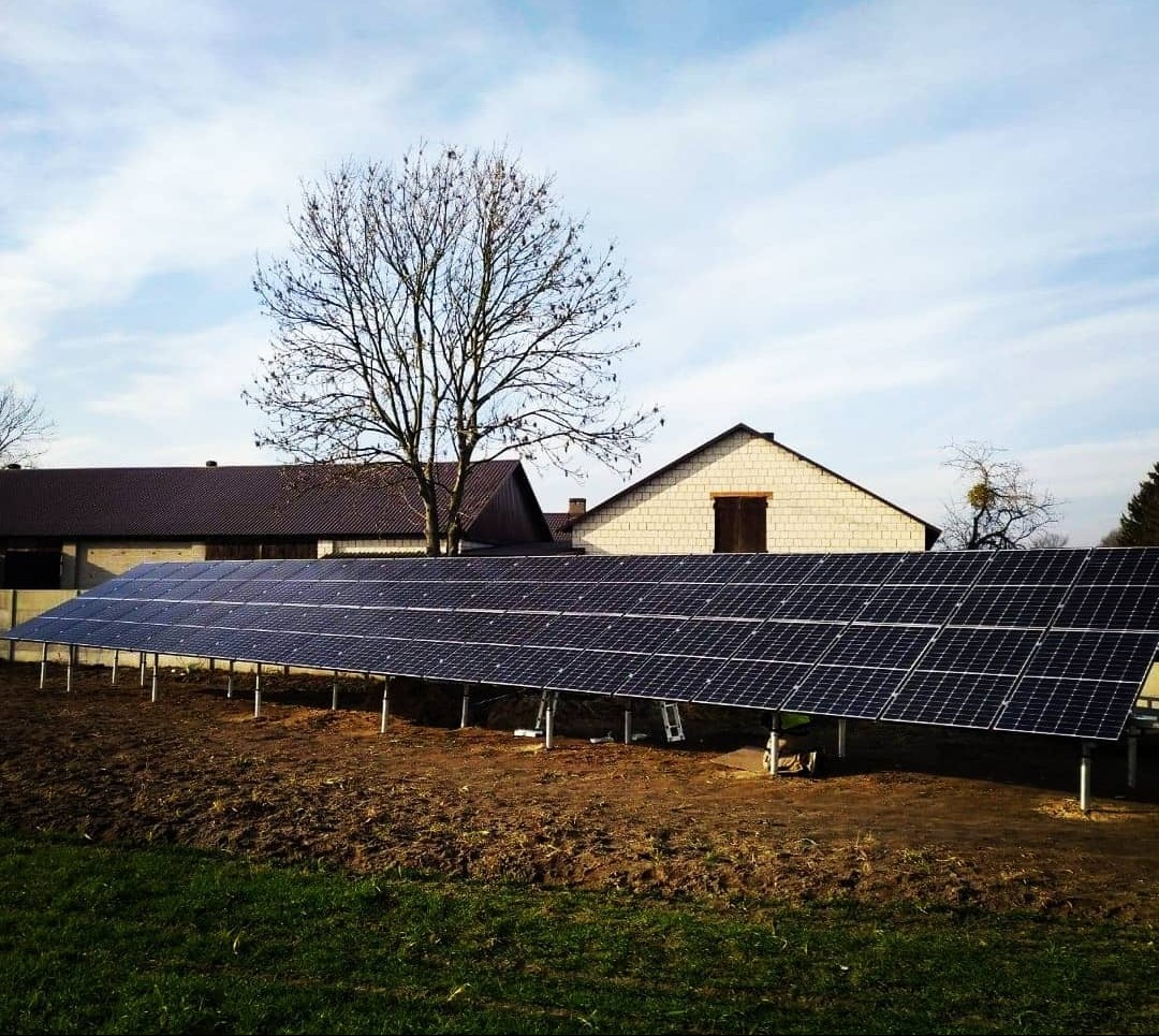 Długa instalacja paneli słonecznych na metalowych podporach, na tle budynków gospodarczych i drzewa bez liści, w słoneczny dzień. Widok z poziomu gruntu.