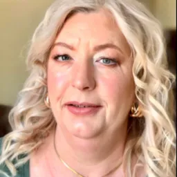 Portret kobiety z blond lokami i delikatnym makijażem, złote kolczyki i naszyjnik z sercem, zielona bluzka, naturalne światło.