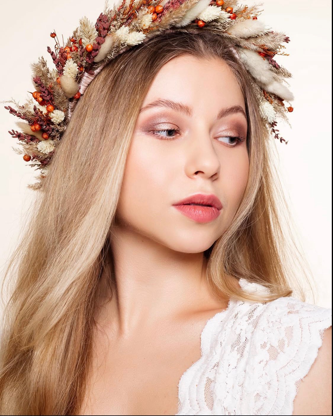 Portret kobiety z blond włosami i kwiatowym wiankiem, subtelny makijaż oczu w odcieniach brązu, delikatne usta, biała koronkowa bluzka, jasne tło.