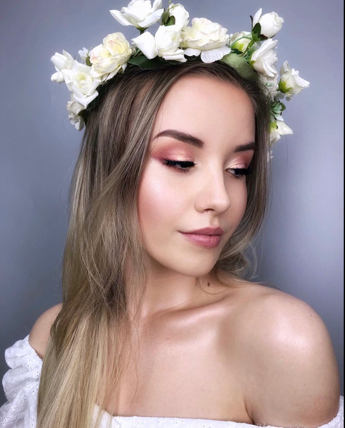 Portret kobiety z kwiatowym wiankiem i delikatnym makijażem w odcieniach różu, z subtelnym rozświetleniem na dekolcie i ramionach, na gładkim, szarym tle.