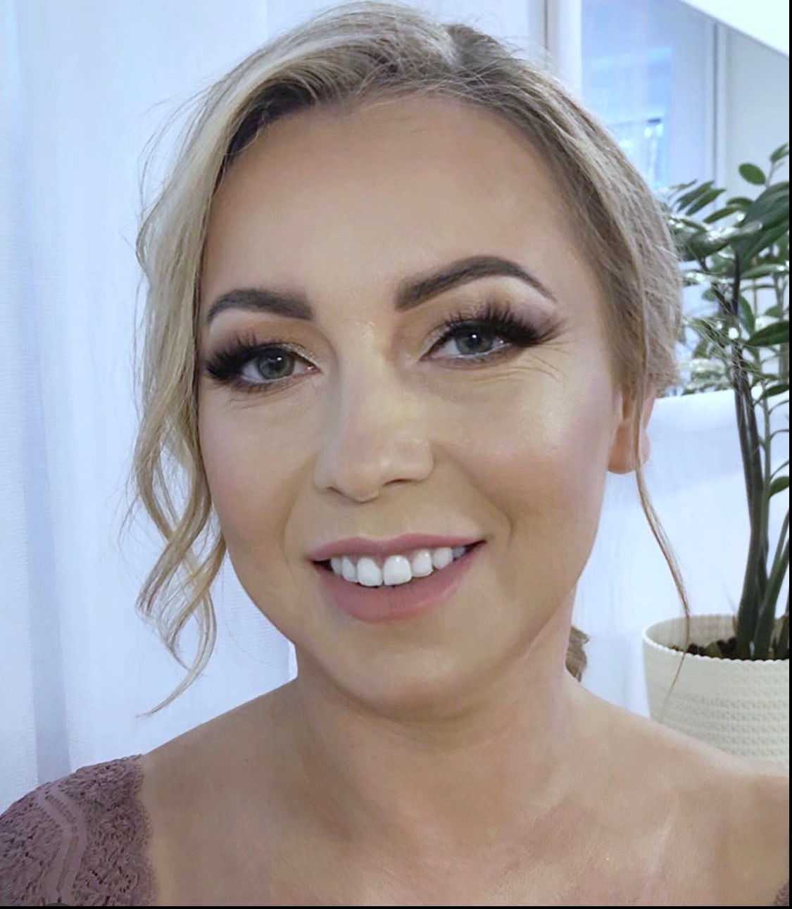 Uśmiechnięta blondynka z profesjonalnym makijażem, widoczne precyzyjnie podkreślone oczy i usta w odcieniu nude. W tle biała zasłona i roślina doniczkowa.