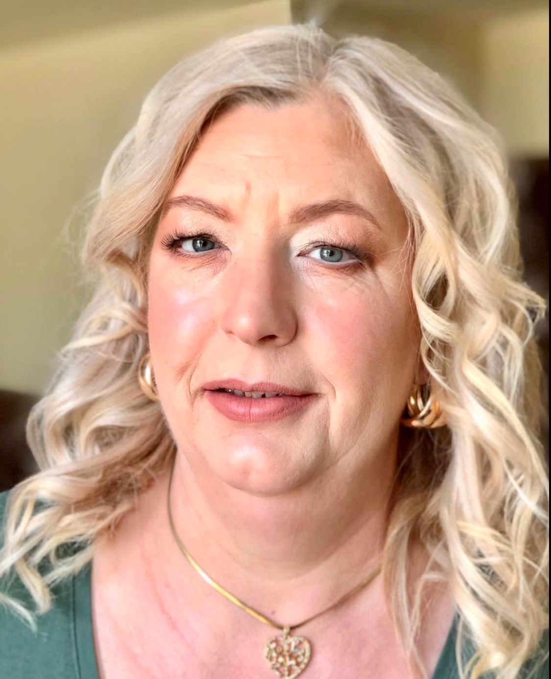 Portret kobiety z blond lokami i delikatnym makijażem, złote kolczyki i naszyjnik z sercem, zielona bluzka, naturalne światło.