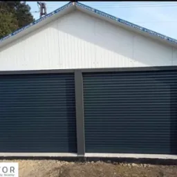 Nowoczesny garaż z dwoma szarymi bramami rolowanymi, białą elewacją i szarym obramowaniem, widoczny logotyp producenta na dole po lewej stronie.