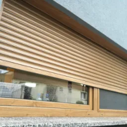 Drewnopodobna roleta zewnętrzna częściowo opuszczona, z widocznym oknem i fragmentem moskitiery w dolnej części ramy okiennej, na tle szarej elewacji.