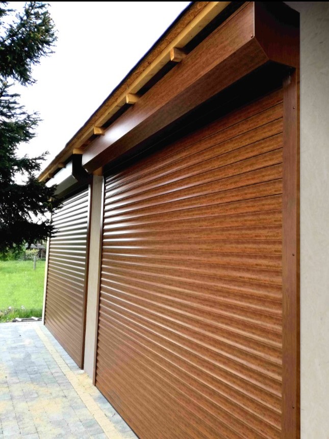 Dwie brązowe rolety zewnętrzne zamontowane na budynku, widok z boku, z widoczną kostką brukową i fragmentem trawnika w tle.