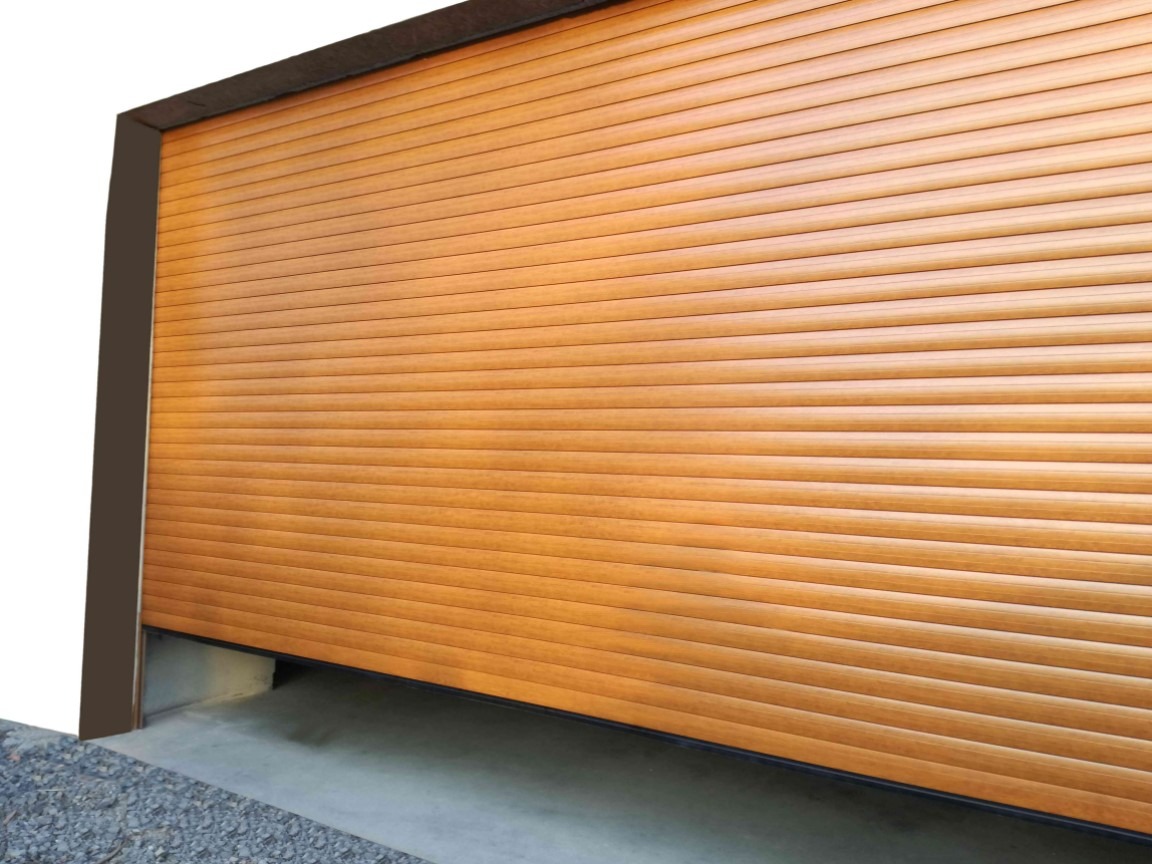 Zamykana brama garażowa segmentowa w kolorze jasnego drewna, częściowo opuszczona, z widocznym fragmentem posadzki i szarego żwiru przed bramą.