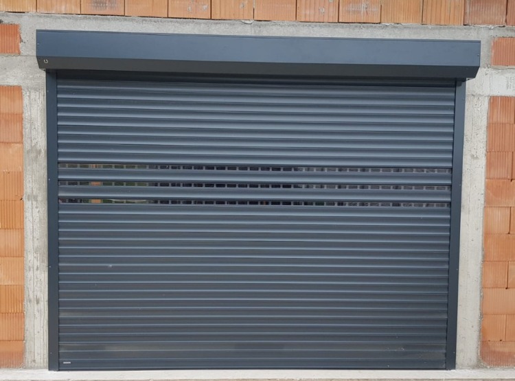 Grafitowa roleta zewnętrzna z perforowanymi lamelami, zamontowana w oknie w surowym stanie, z widocznym murem z cegły i betonowym nadprożem.