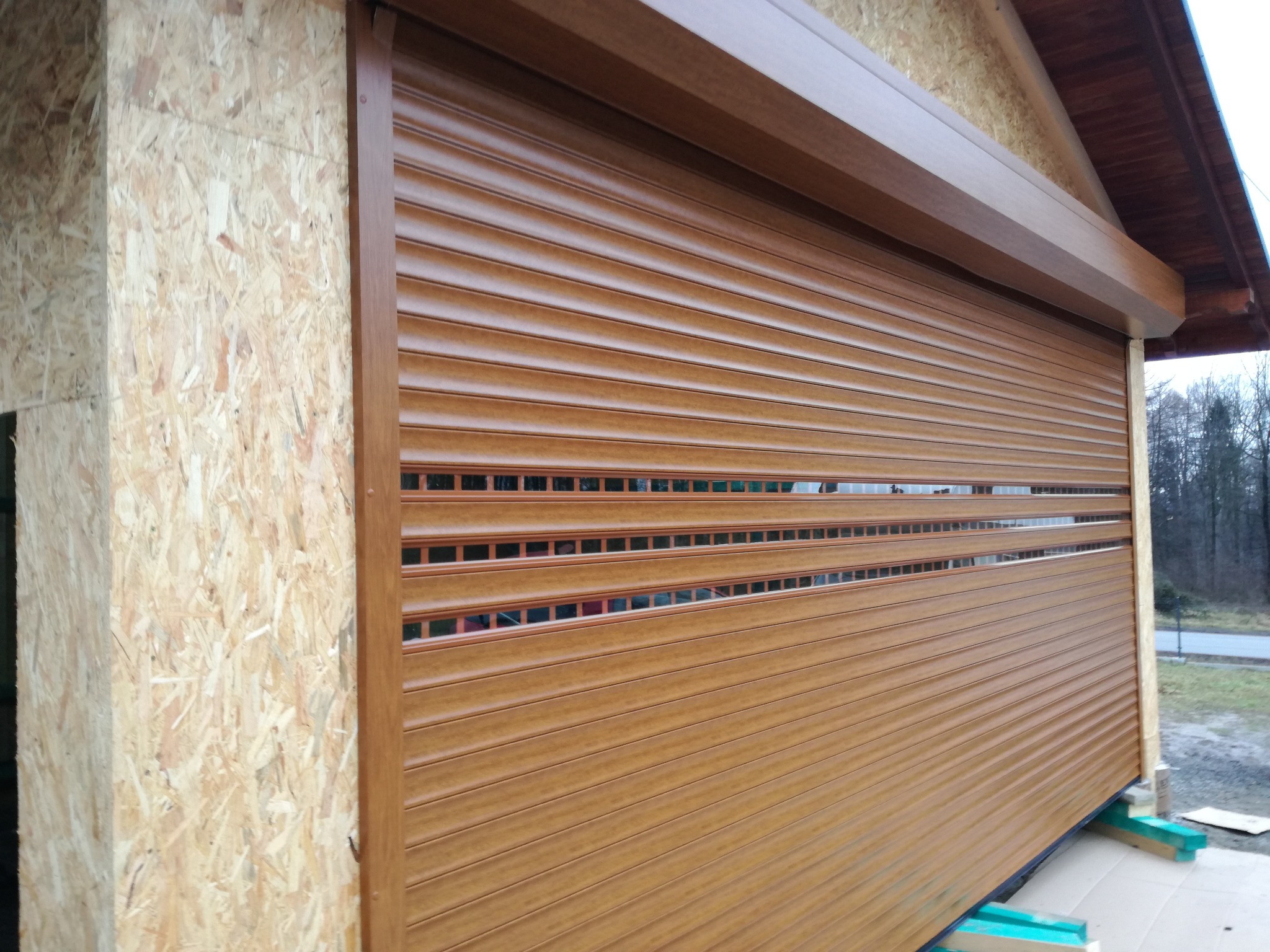 Brązowa roleta zewnętrzna częściowo opuszczona, widoczna na tle surowej konstrukcji budynku z płyty OSB i elementami drewnianymi, z prześwitami ukazującymi otoczenie.
