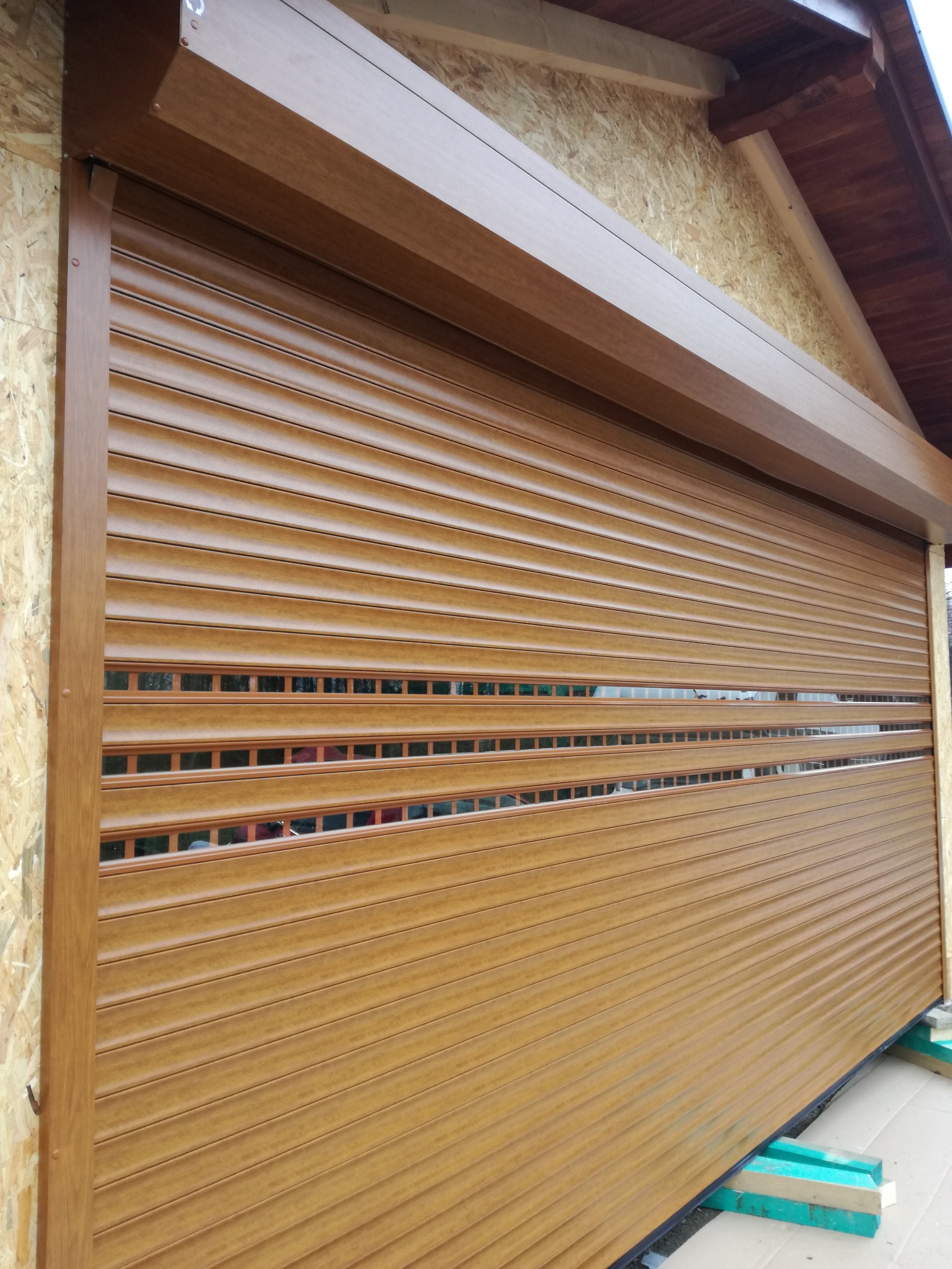 Zamknięta, brązowa roleta zewnętrzna z poziomymi lamelami, zamontowana na elewacji domu z widoczną konstrukcją dachu i fragmentem płyty OSB pod okapem.