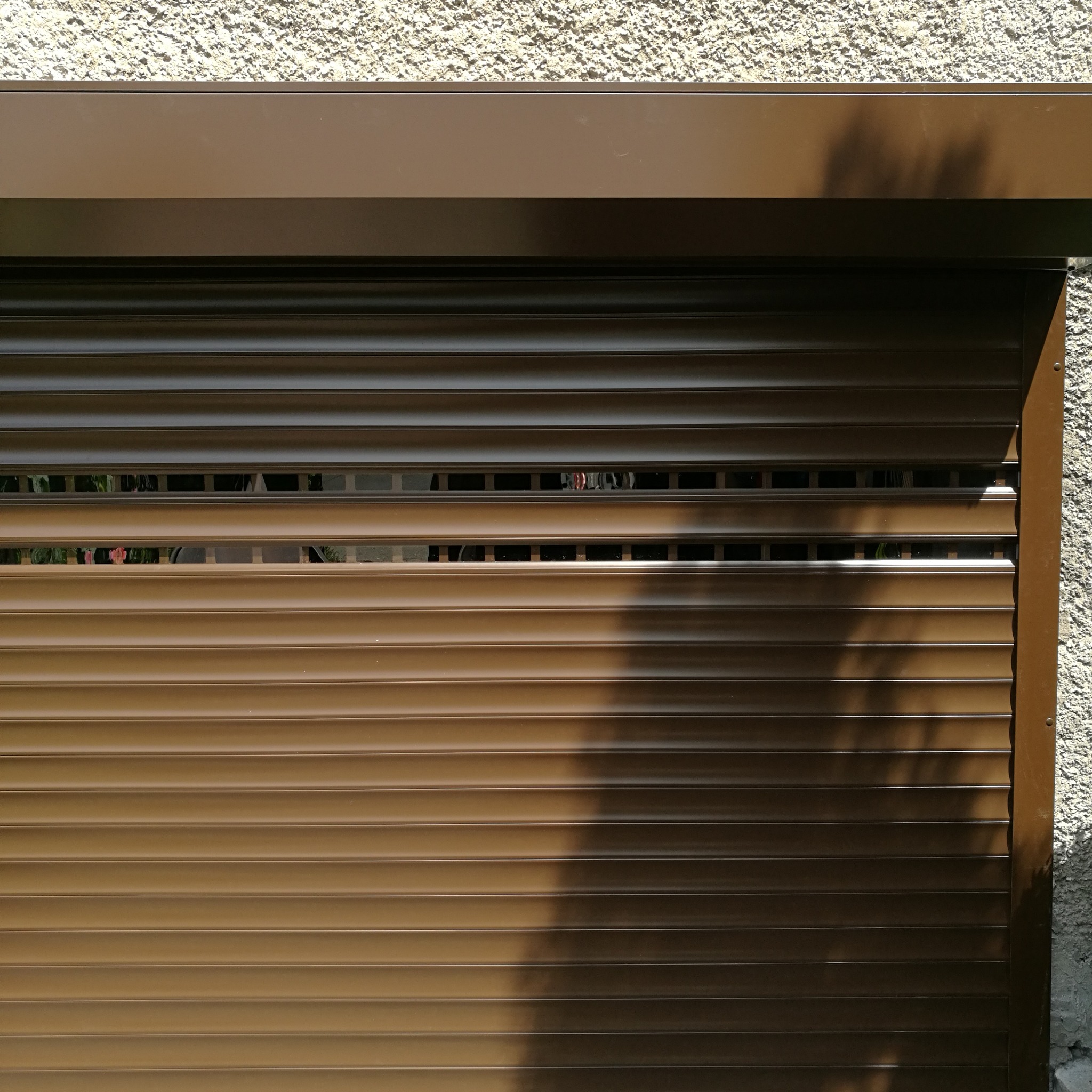 Częściowo opuszczona, brązowa roleta zewnętrzna z widocznym fragmentem elewacji o szorstkiej fakturze i cieniami drzew.