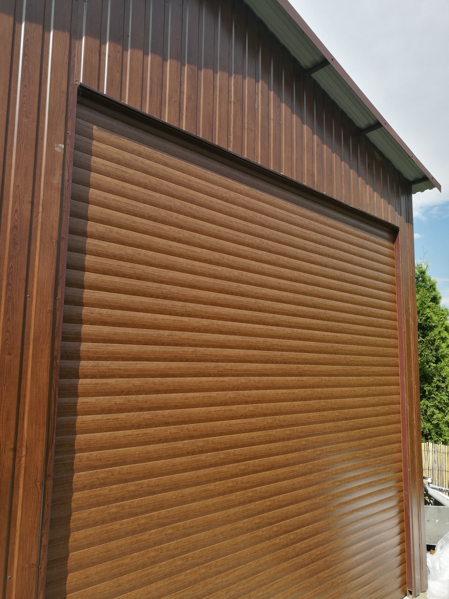 Zewnętrzna, brązowa roleta na budynku z blachy trapezowej, widok z dołu na zamkniętą konstrukcję, z fragmentem nieba i zieleni w tle.