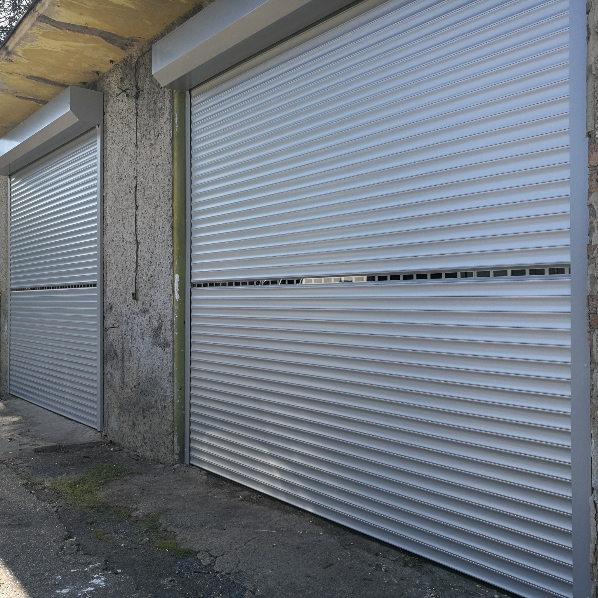 Dwie szare rolety zewnętrzne zamontowane na szorstkiej, betonowej ścianie budynku, jedna z nich lekko podniesiona, odsłaniając fragment wnętrza.