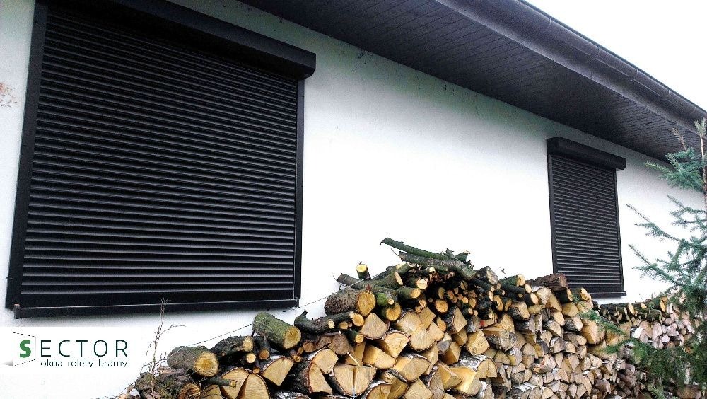 Elewacja domu z dwoma oknami wyposażonymi w czarne rolety zewnętrzne, ułożone drewno opałowe wzdłuż ściany, widoczne logo firmy 'Sector' w lewym dolnym rogu.