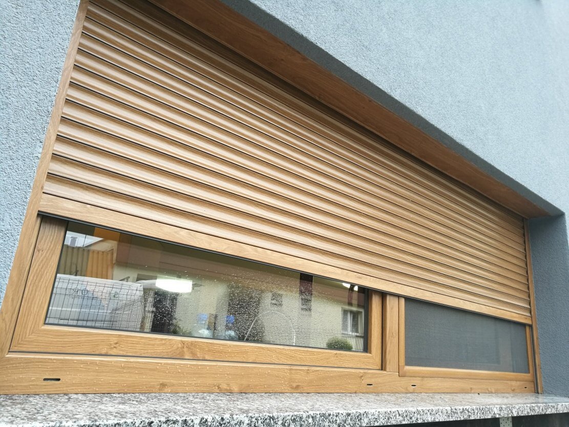 Drewnopodobna roleta zewnętrzna częściowo opuszczona, z widocznym oknem i fragmentem moskitiery w dolnej części ramy okiennej, na tle szarej elewacji.