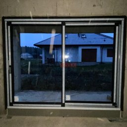 LASERVICE-OKNA - Duże przesuwne okno z czarną ramą w surowym stanie, widok na dom z dachem pokrytym szarą dachówką w tle.