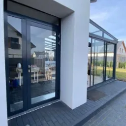 Nowoczesne, grafitowe drzwi tarasowe z widokiem na wnętrze z meblami ogrodowymi i przeszklony ogród zimowy, kostka brukowa na zewnątrz.