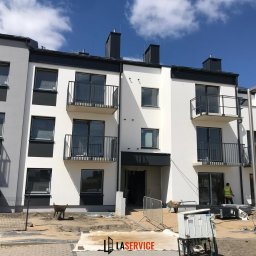 LASERVICE SPÓŁKA Z OGRANICZONĄ ODPOWIEDZIALNOŚCIĄ - Nowoczesny budynek wielorodzinny z balkonami i oknami, w trakcie wykańczania. Widoczni pracownicy i materiały budowlane. Dzień słoneczny, błękitne niebo.