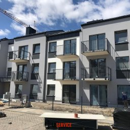 LASERVICE SPÓŁKA Z OGRANICZONĄ ODPOWIEDZIALNOŚCIĄ - Nowoczesny budynek wielorodzinny w szaro-białej kolorystyce z balkonami, widok z zewnątrz. Na pierwszym planie materiały budowlane i robotnik w oknie.