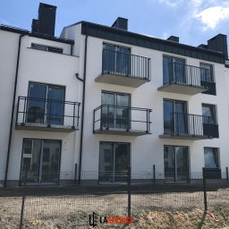 LASERVICE-OKNA - Nowoczesny budynek wielorodzinny z białymi ścianami, czarnymi ramami okien i balkonami z metalowymi balustradami. Widok z zewnątrz, ogrodzenie tymczasowe.