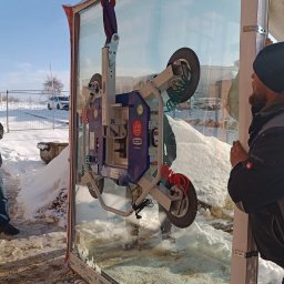 LASERVICE SPÓŁKA Z OGRANICZONĄ ODPOWIEDZIALNOŚCIĄ - Montaż okna z użyciem przyssawki próżniowej na budowie w zimowych warunkach. Widoczny pracownik i fragment krajobrazu z białym samochodem w tle.