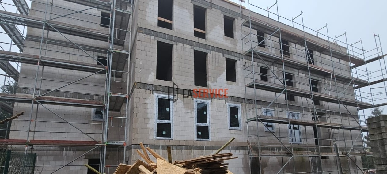 Budynek w trakcie budowy z częściowo zamontowanymi oknami, otoczony rusztowaniami i stertą płyt OSB, widoczne otwory okienne bez okien.