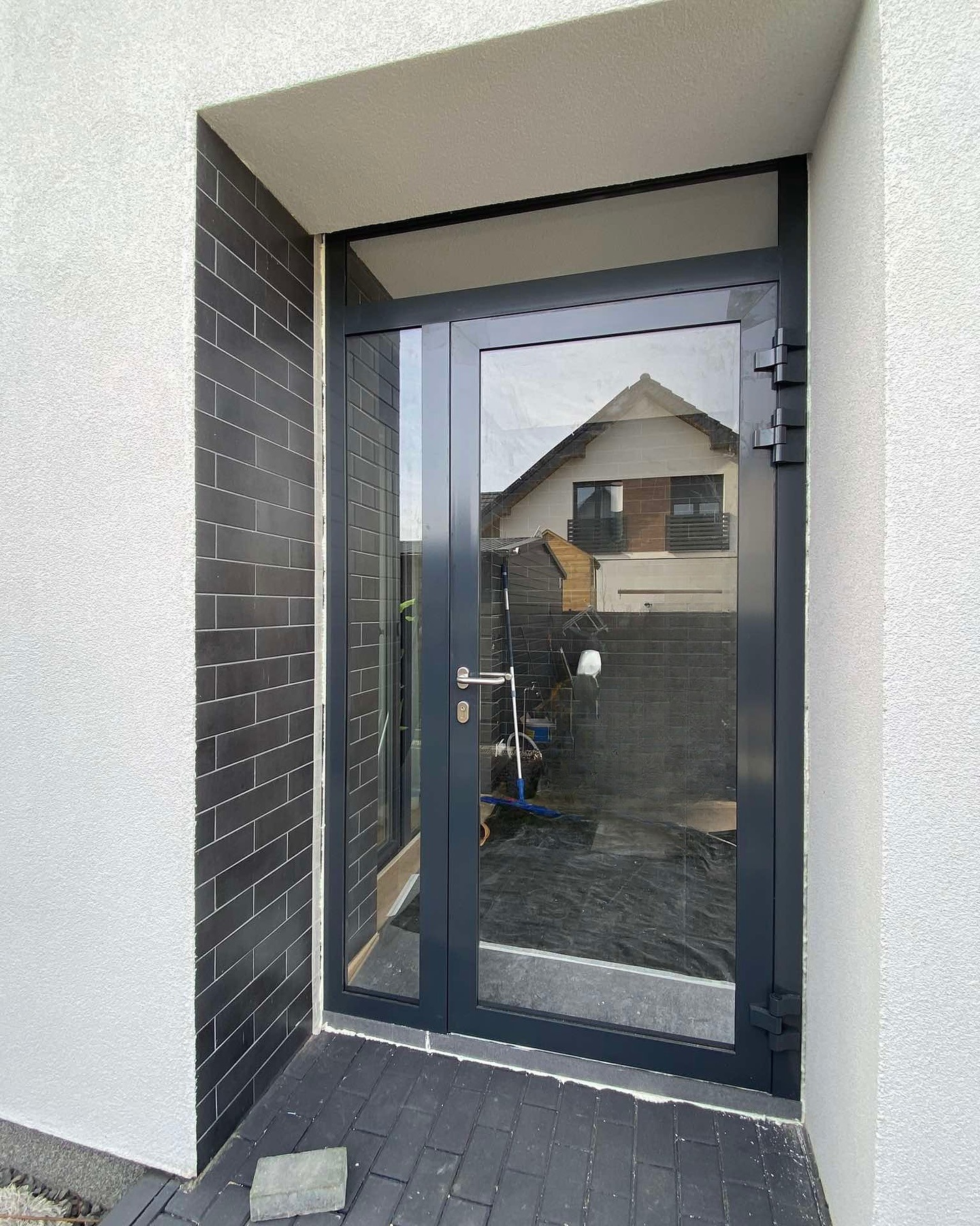 Zewnętrzne drzwi aluminiowe z dużym przeszkleniem, szara rama, widoczne zawiasy, elewacja z białego tynku i czarnej cegły, odbicie budynku w szybie.