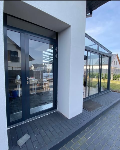 Nowoczesne, grafitowe drzwi tarasowe z widokiem na wnętrze z meblami ogrodowymi i przeszklony ogród zimowy, kostka brukowa na zewnątrz.