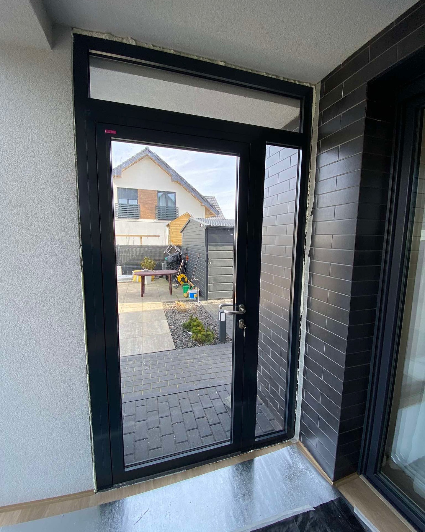 Czarne drzwi wejściowe z przeszkleniami i widokiem na podwórko, zamontowane w domu z szarymi cegłami na ścianie i pianką montażową wokół ramy.