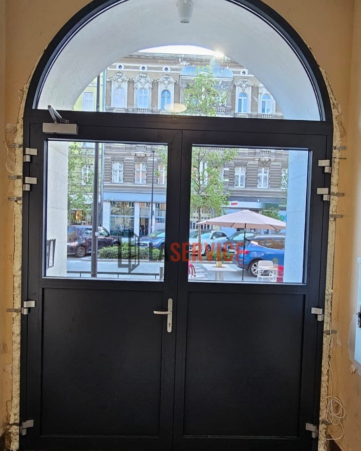 Czarne, dwuskrzydłowe drzwi z przeszkleniami, w łukowym otworze. Widoczne elementy montażowe i fragment ulicy z budynkami w tle.