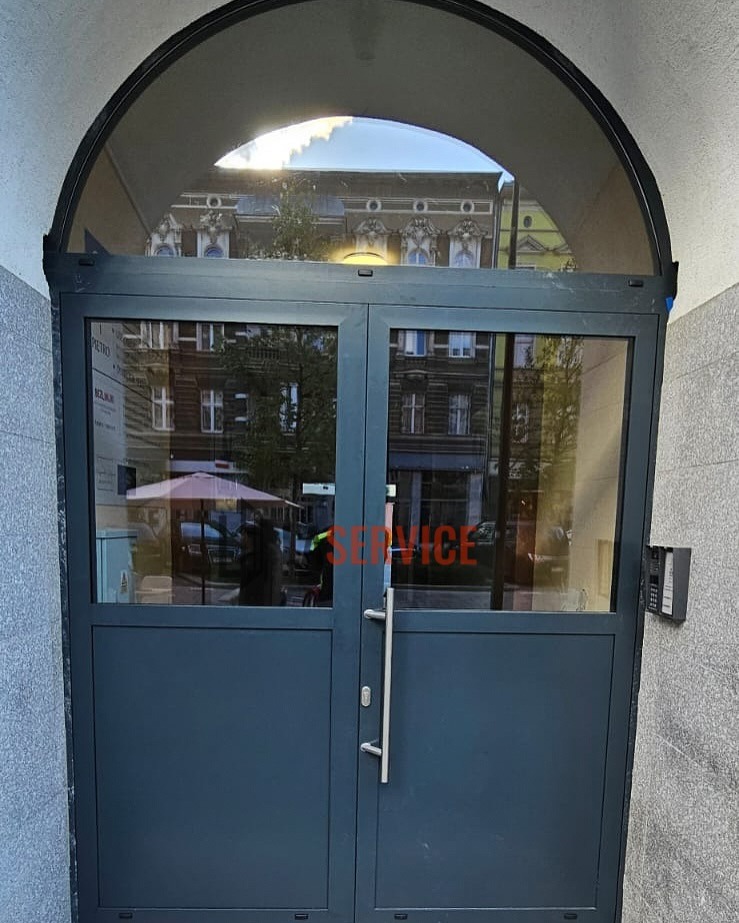 Szare, dwuskrzydłowe drzwi z przeszkleniami pod łukiem, widoczne z zewnątrz. Na szybie odbija się budynek. Metalowa klamka i domofon po prawej stronie.