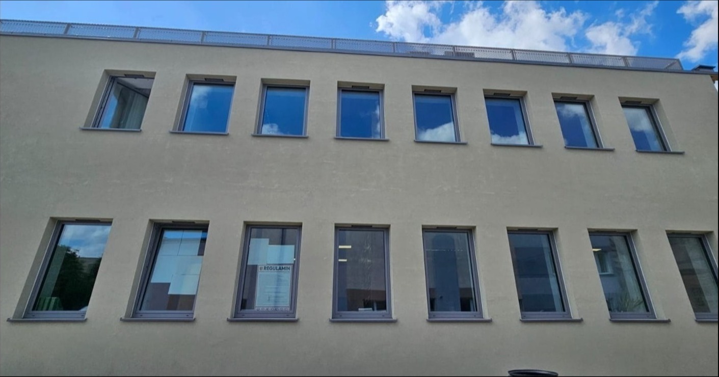 Elewacja budynku z rzędem okien PCV w Szczecinie. W oknach odbija się błękitne niebo z chmurami. Jedno okno z plakatem informacyjnym.