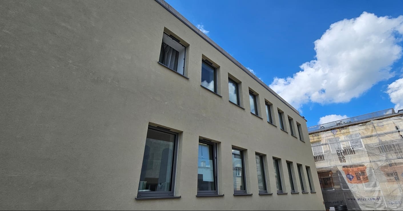 Elewacja budynku z zamontowanymi oknami. Widoczne szare ramy okienne i błękitne niebo z chmurami w tle. Ujęcie z dołu, podkreślające regularny układ okien.
