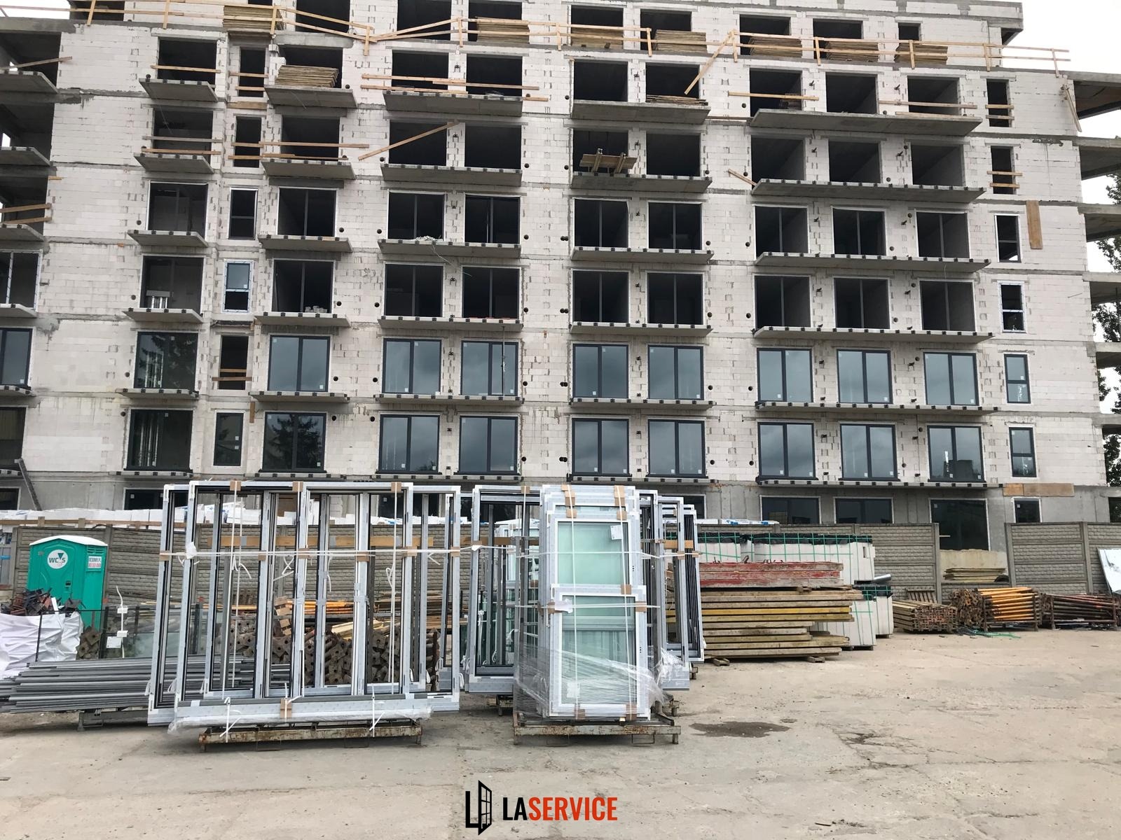 Budynek w trakcie montażu okien. Na pierwszym planie ułożone ramy okienne, w tle surowy budynek z częściowo zamontowanymi oknami. Widoczne rusztowania i materiały budowlane.