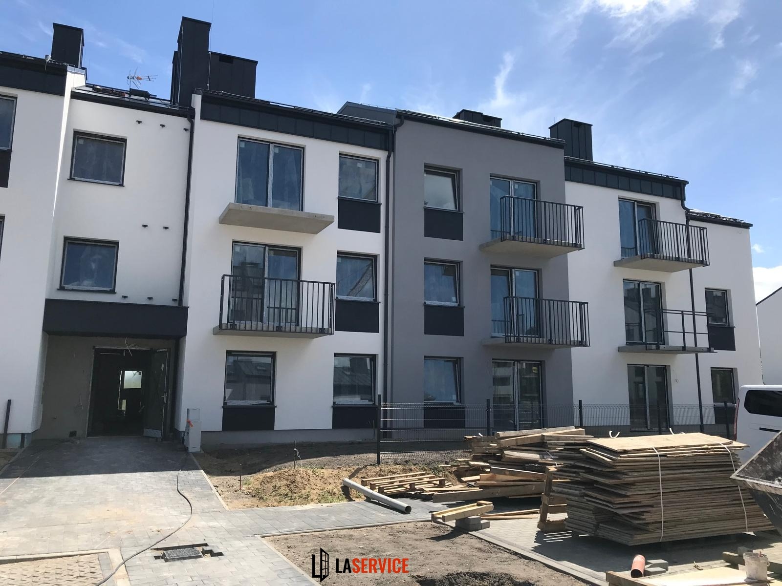 Nowoczesny, trzypiętrowy budynek apartamentowy w biało-szarej kolorystyce, z balkonami z czarną balustradą. Częściowo wykończony teren budowy na pierwszym planie.
