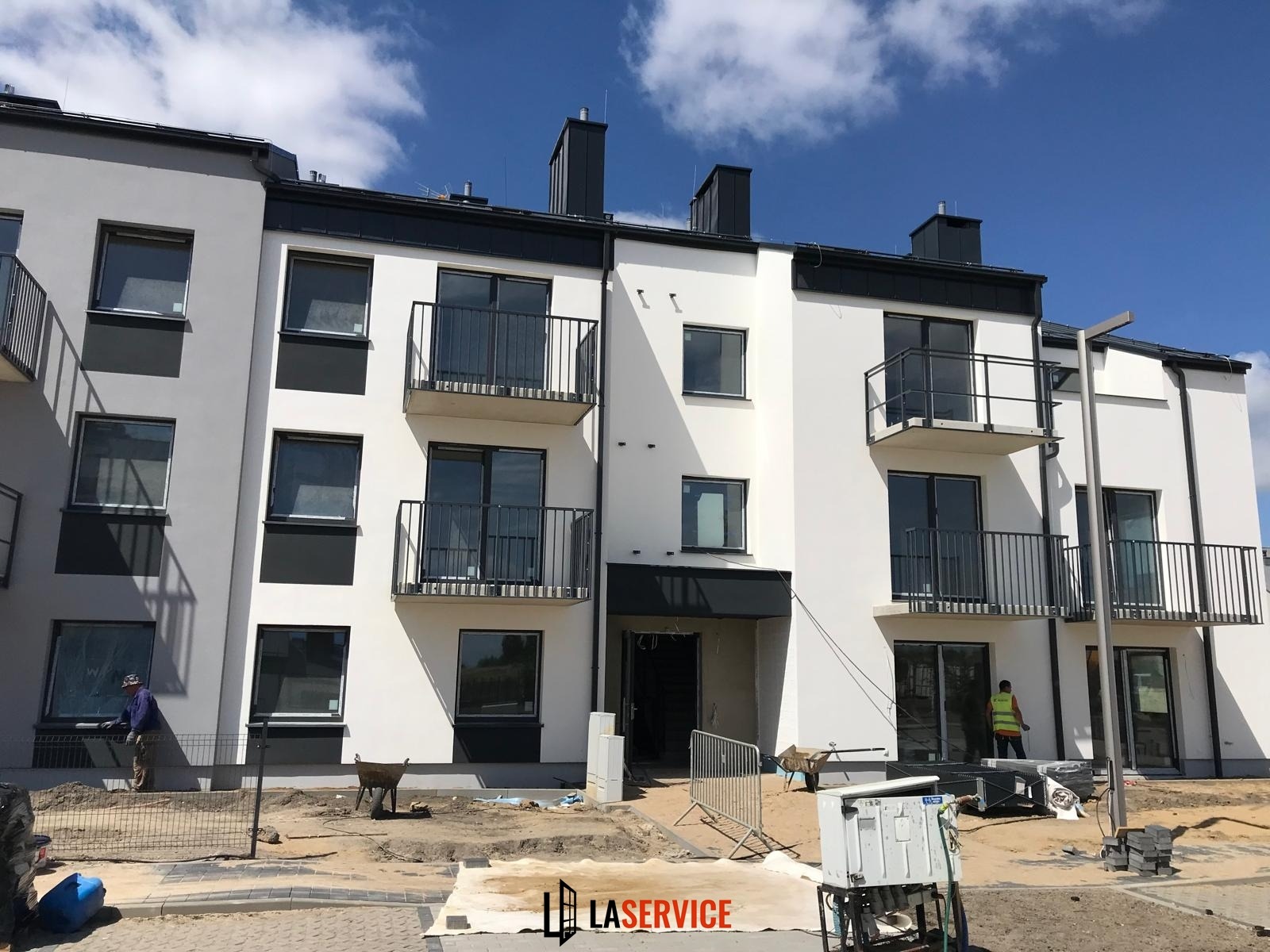Nowoczesny budynek wielorodzinny z balkonami i oknami, w trakcie wykańczania. Widoczni pracownicy i materiały budowlane. Dzień słoneczny, błękitne niebo.