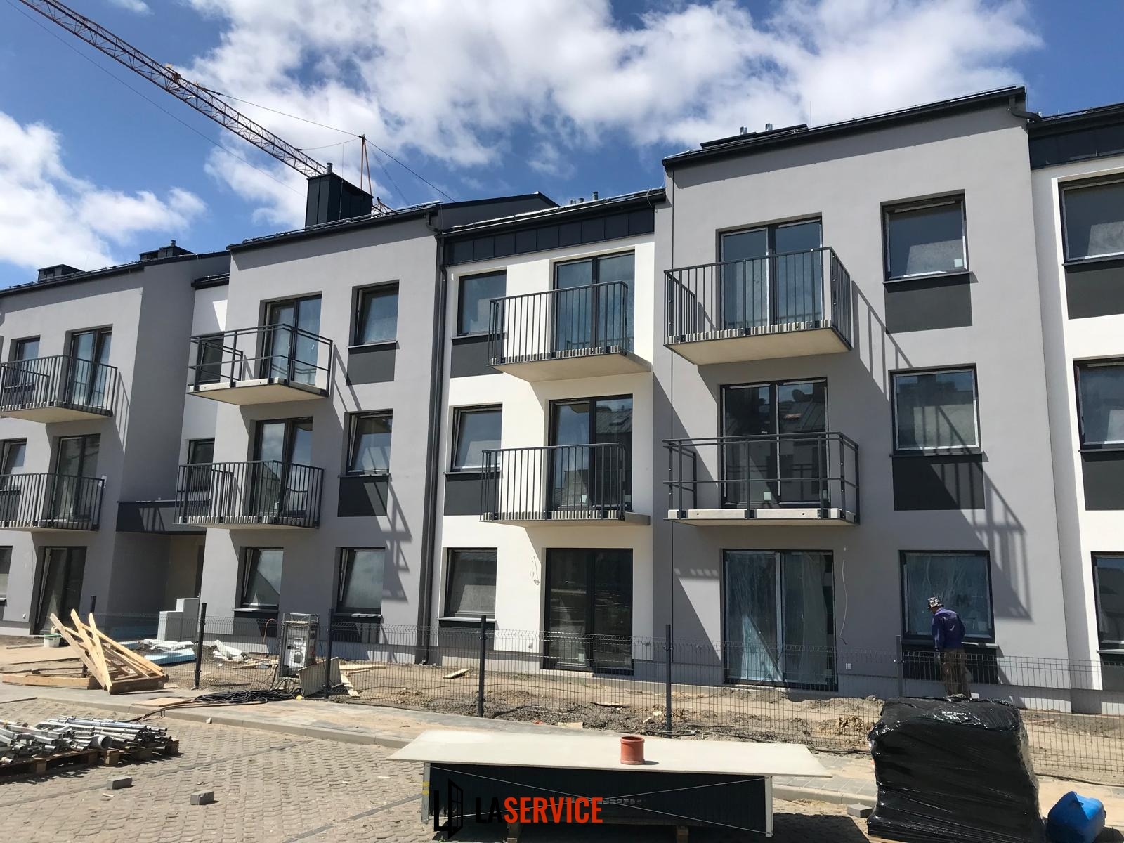 Nowoczesny budynek wielorodzinny w szaro-białej kolorystyce z balkonami, widok z zewnątrz. Na pierwszym planie materiały budowlane i robotnik w oknie.