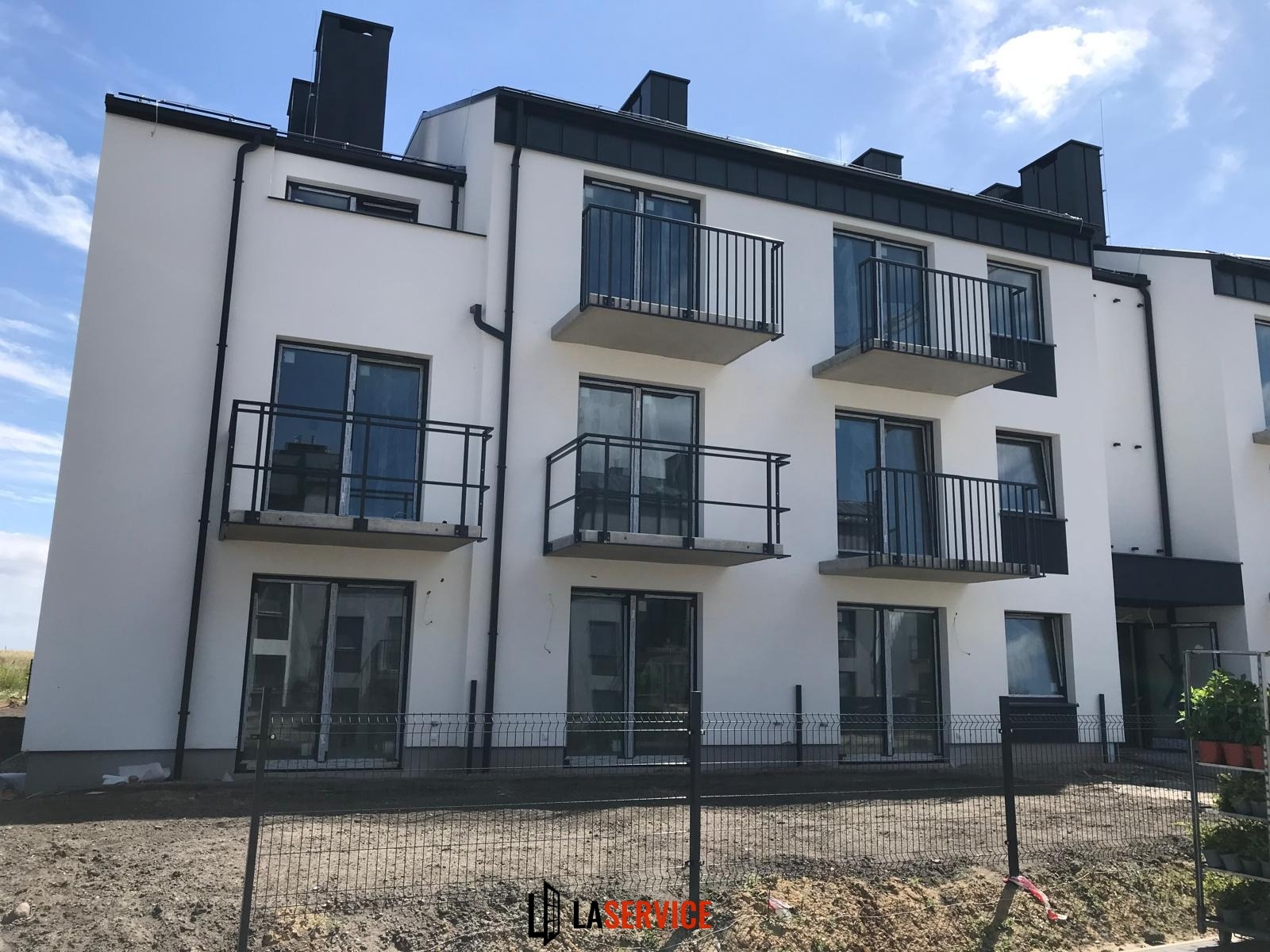 Nowoczesny budynek wielorodzinny z białymi ścianami, czarnymi ramami okien i balkonami z metalowymi balustradami. Widok z zewnątrz, ogrodzenie tymczasowe.