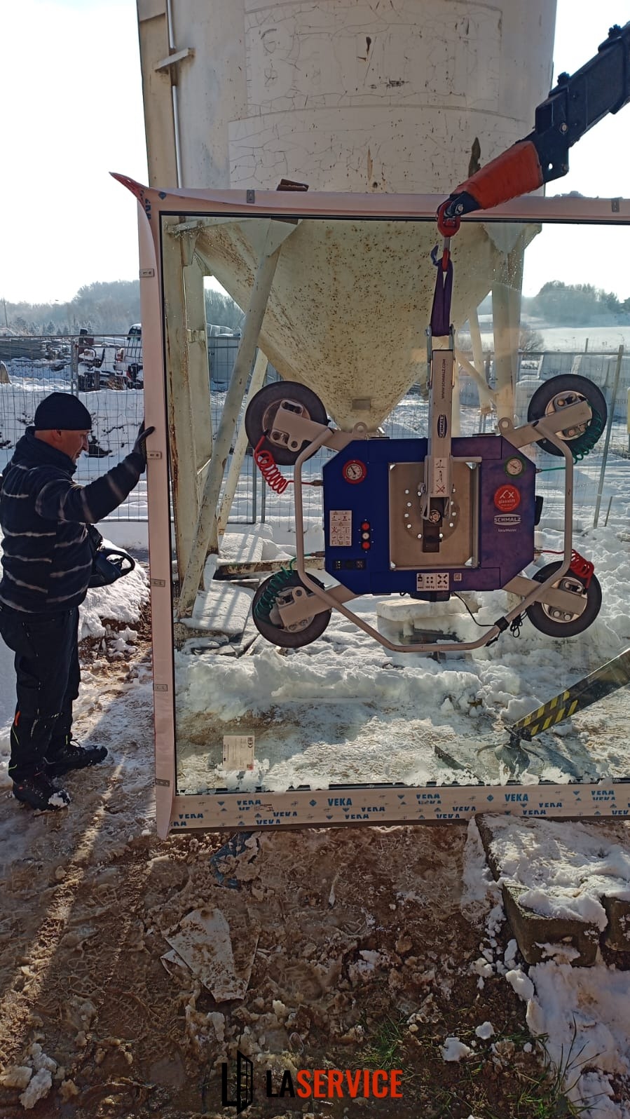 Montera okienny z przyssawką Schmalz EasyMaster przenosi duże okno Veka na tle zimowego krajobrazu i betonowego silosu. Widoczne ślady w śniegu.