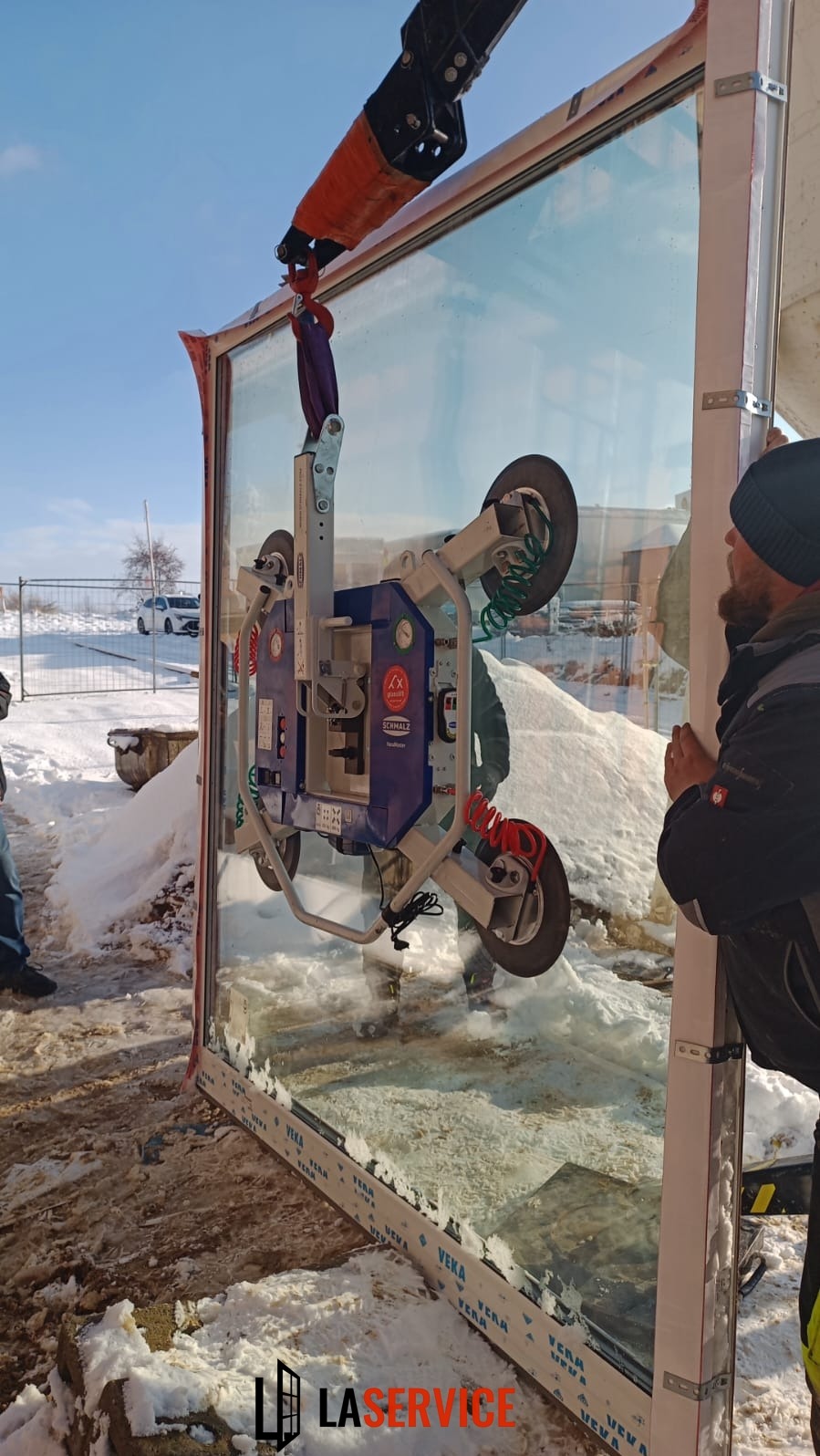 Montaż okna z użyciem przyssawki próżniowej na budowie w zimowych warunkach. Widoczny pracownik i fragment krajobrazu z białym samochodem w tle.