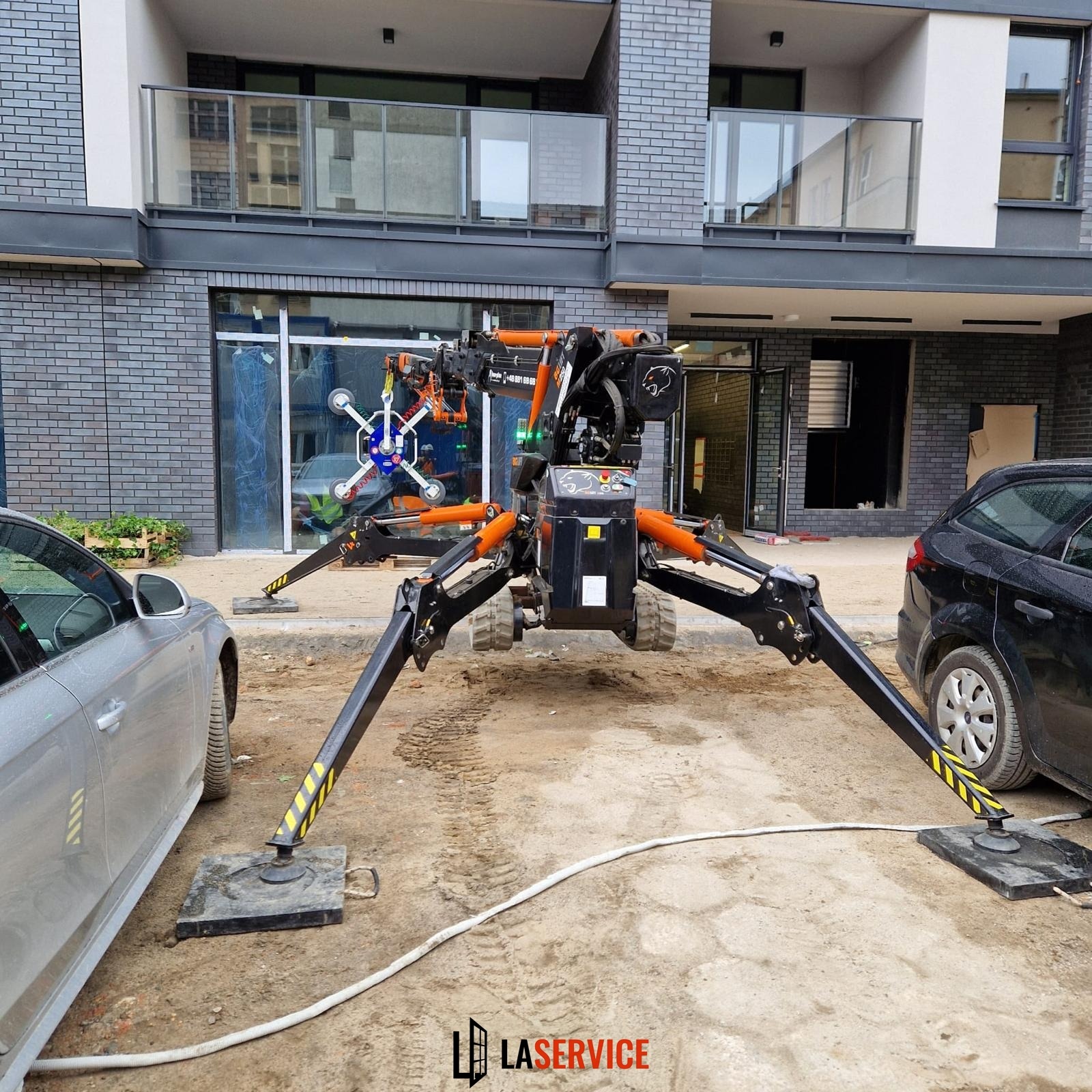 Montaż okna w nowym budynku za pomocą specjalistycznego dźwigu ssącego. Widoczne mocowania przyssawkowe na szybie i czarno-pomarańczowa konstrukcja urządzenia.