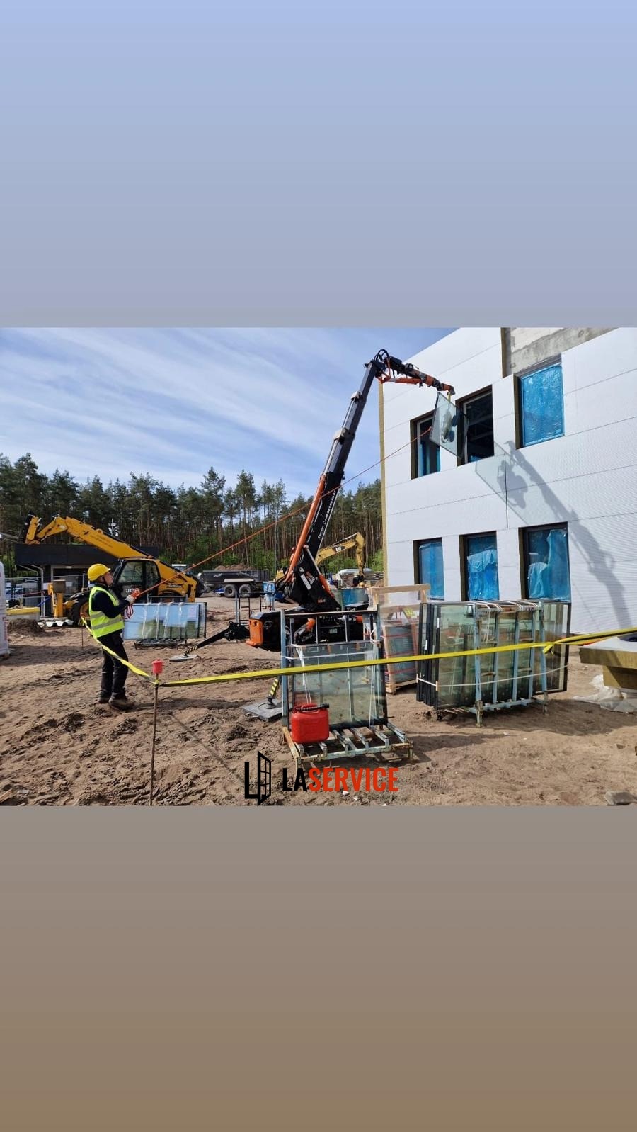 Montaż okien na budowie: pracownik w kamizelce nadzoruje dźwig wstawiający okno w otwór, z boku stoją okna zabezpieczone folią, widoczne maszyny budowlane.