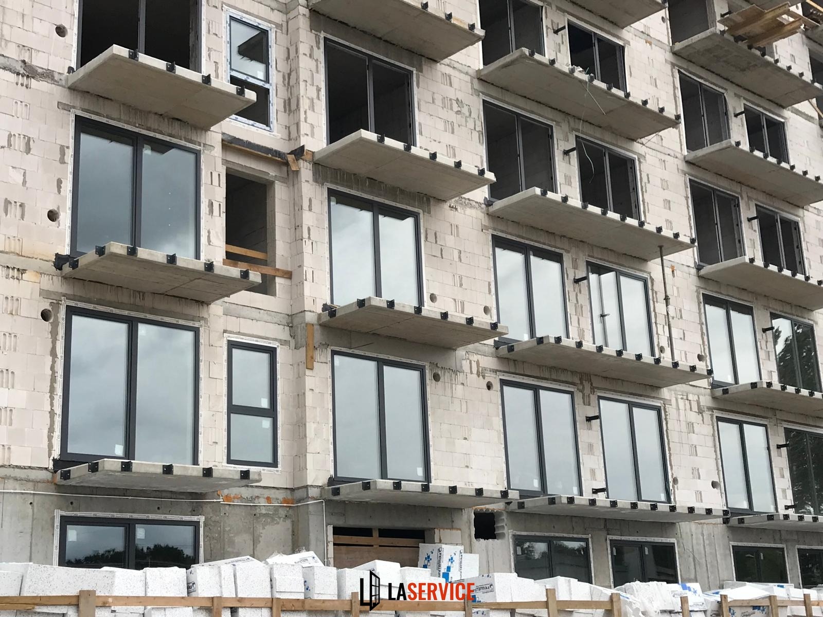 Montaż czarnych ram okiennych w surowym budynku z betonu komórkowego. Część okien jeszcze bez szyb. Widoczne materiały budowlane.