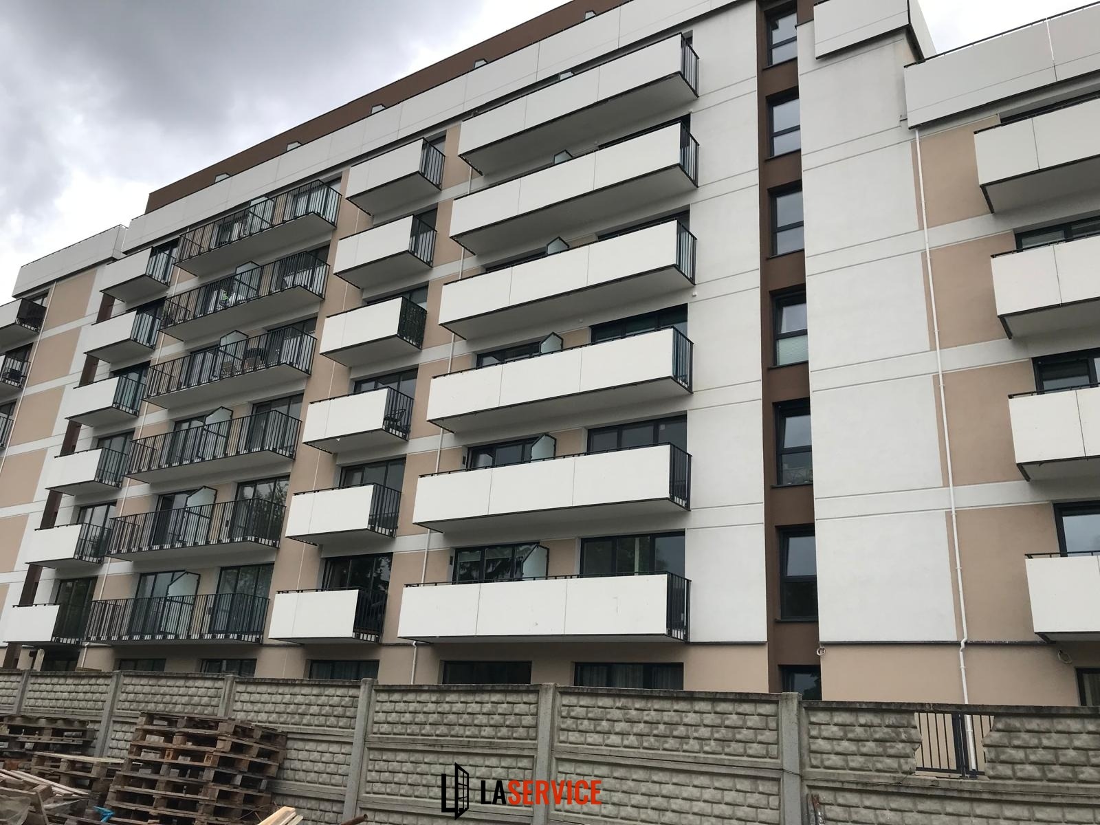 Fasada budynku wielorodzinnego z balkonami o białych balustradach i czarnych barierkach, widziana z dołu, z paletami i betonowym ogrodzeniem na pierwszym planie.