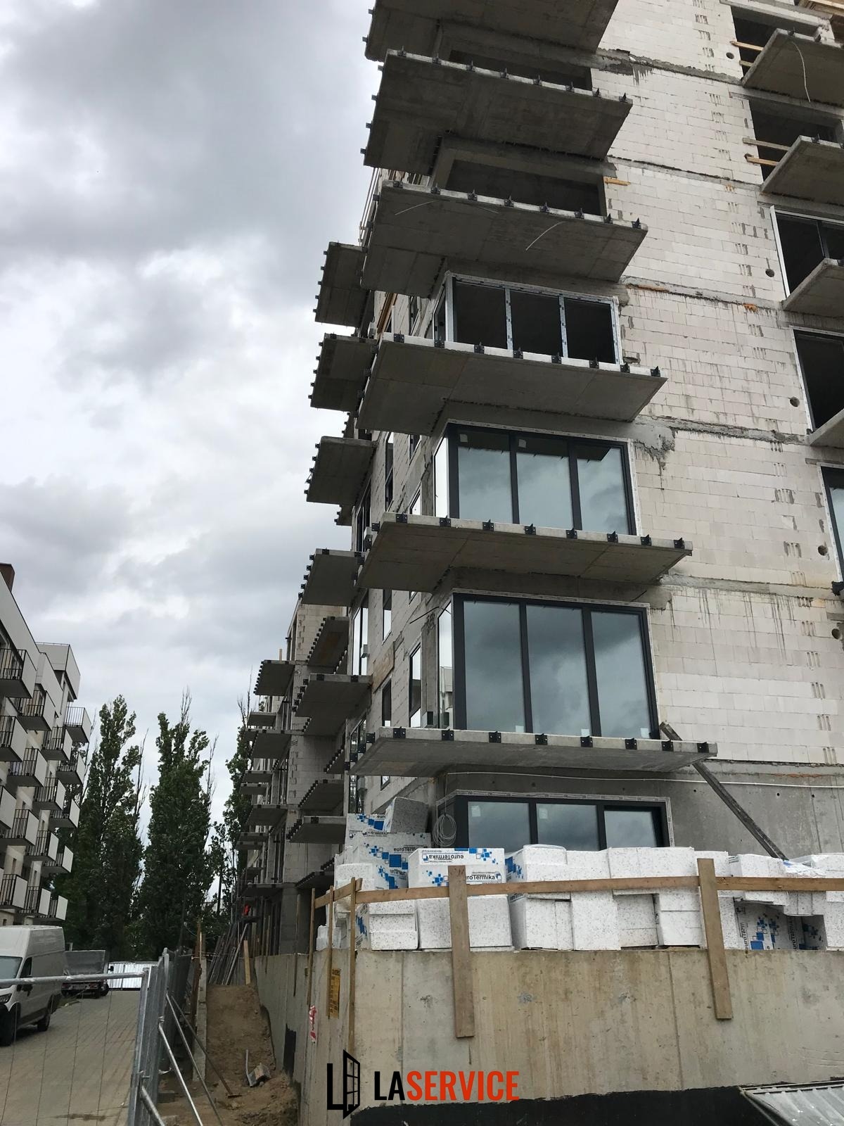 Budynek w trakcie budowy z zamontowanymi oknami na kilku piętrach. Widoczne materiały budowlane i surowe wykończenie. Pochmurne niebo w tle.