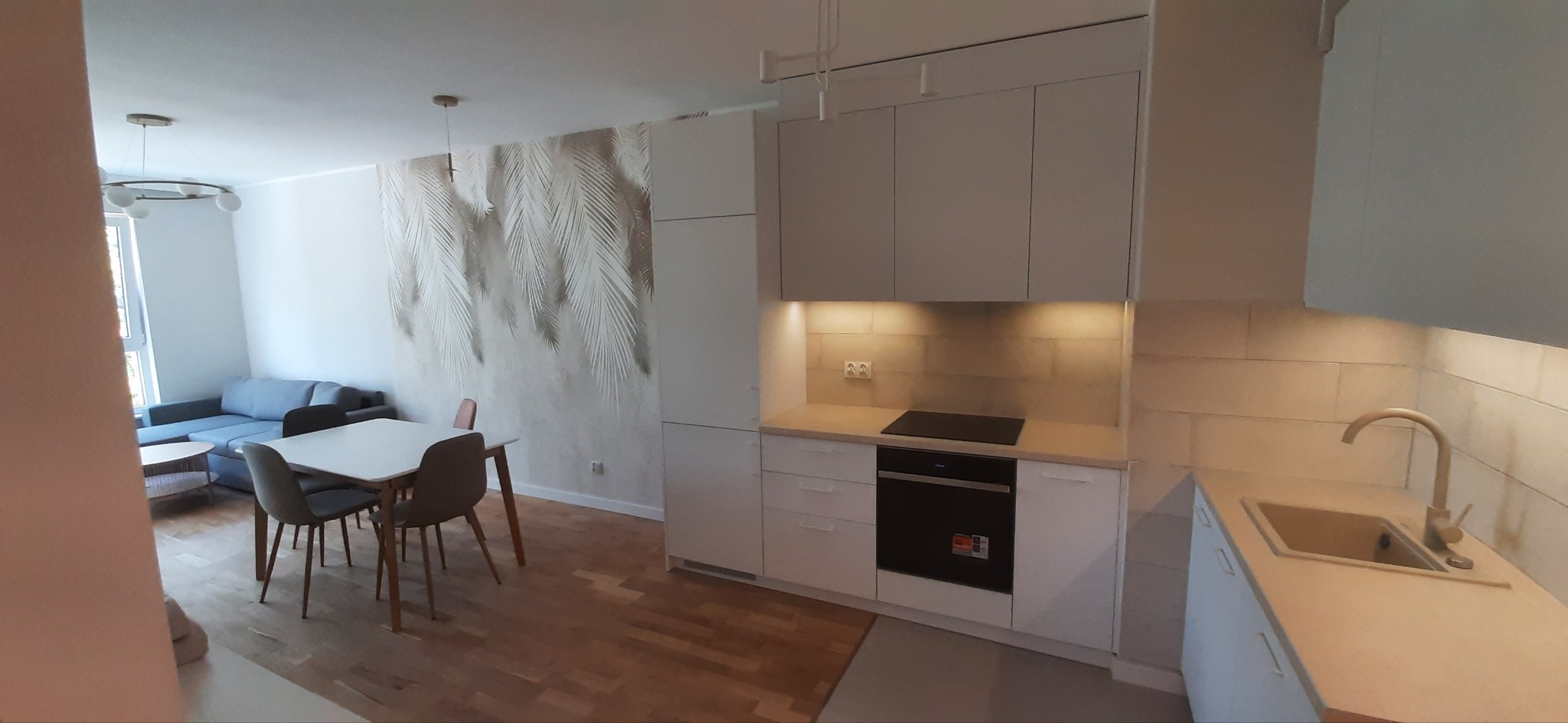 Jasna kuchnia z zabudową na wymiar, widok na salon z szarą sofą, na ścianie tapeta z motywem liści palmowych.