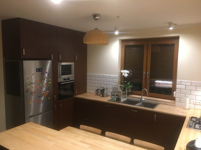 Ciemnobrązowa zabudowa kuchenna z drewnianym blatem, białymi płytkami na ścianie i stalową lodówką ozdobioną magnesami. W oknie widoczny kwiat orchidei.